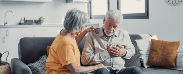 Older white woman on couch comforting older white man who holds his chest 709688414